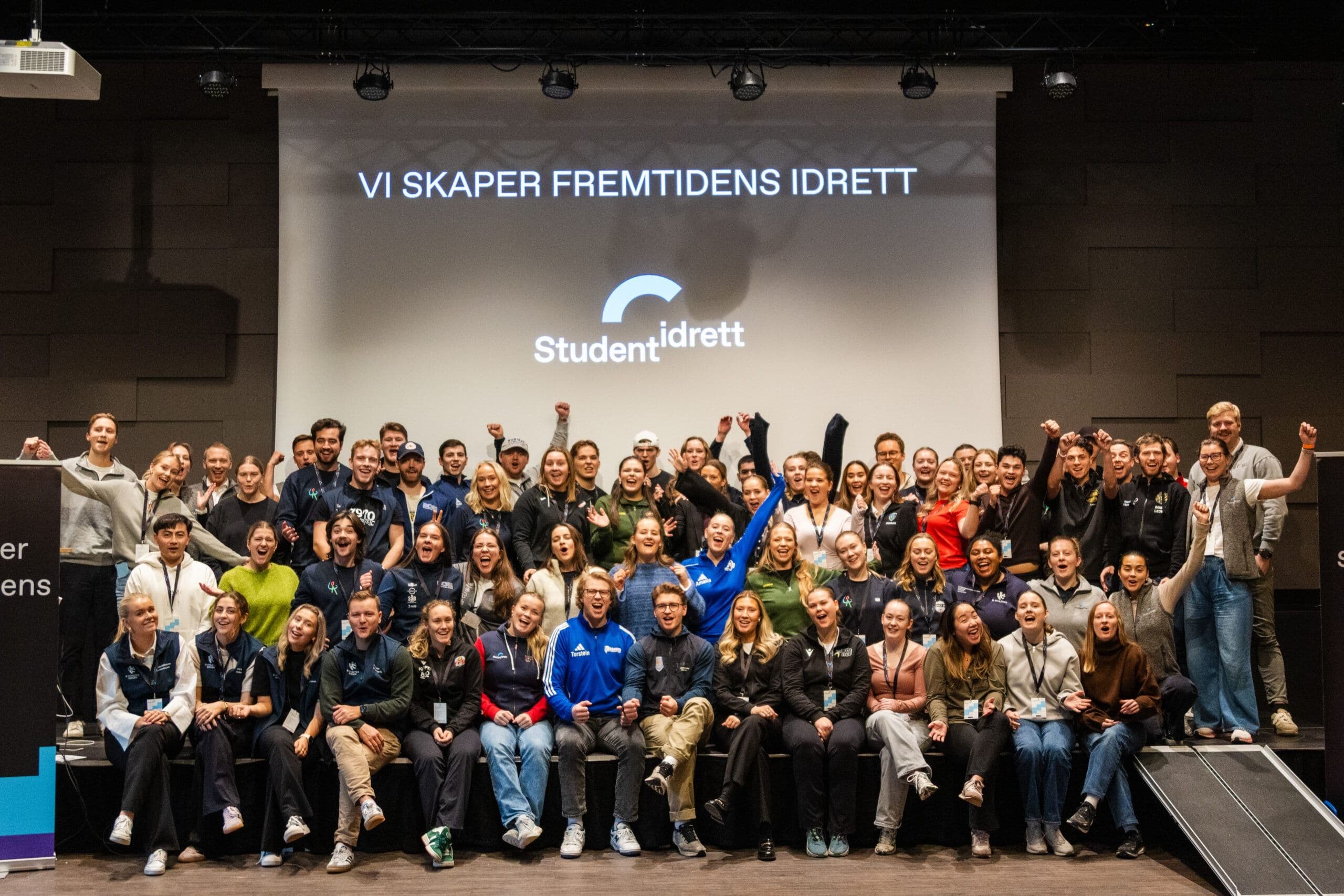 En stor gruppe studenter poserer jublende på en scene foran en skjerm med teksten Vi skaper fremtidens idrett og Studentidrett-logoen.
