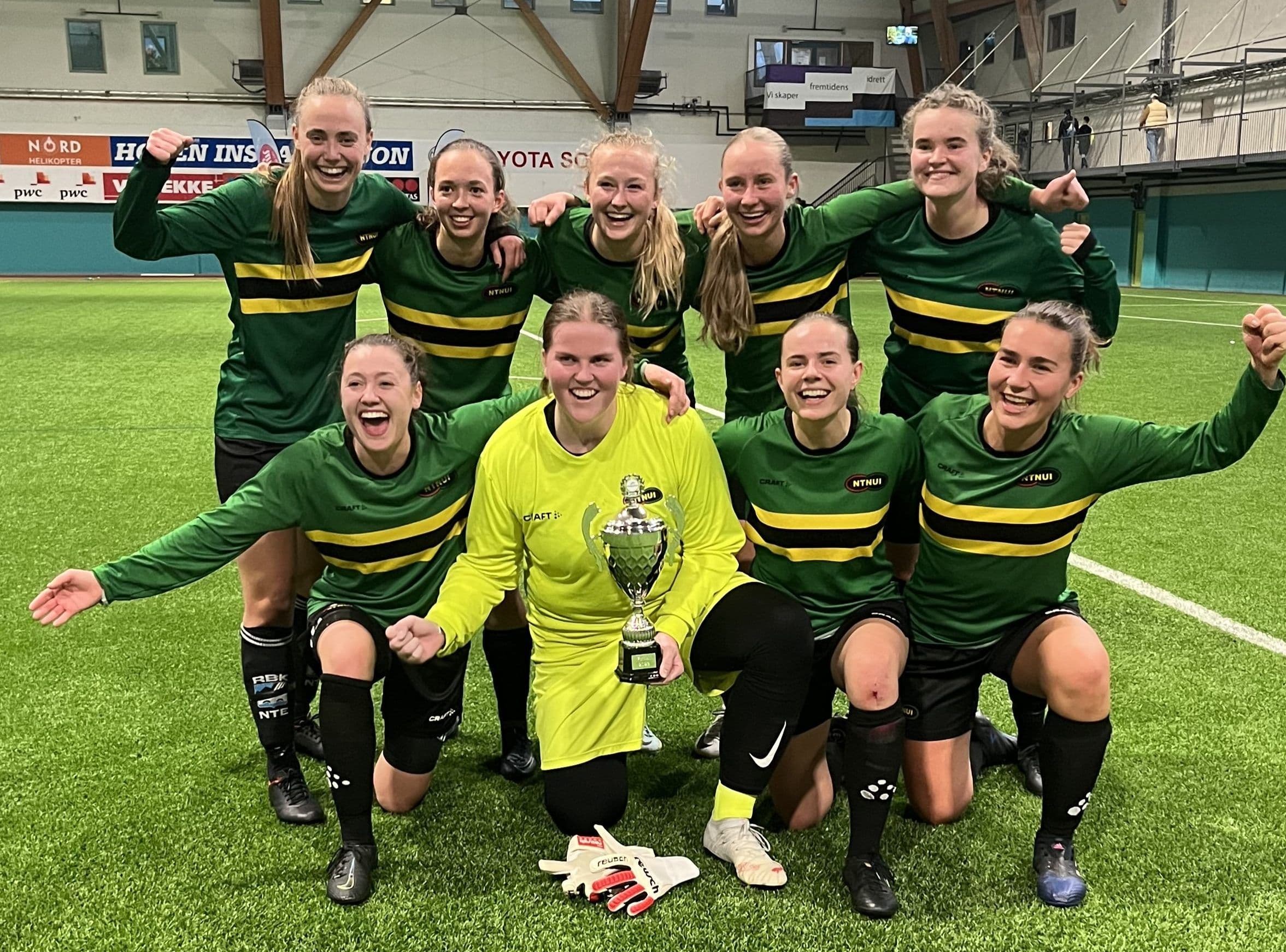 Et jublende kvinnelag i grønne og gule drakter poserer med en pokal på en innendørs fotballbane.