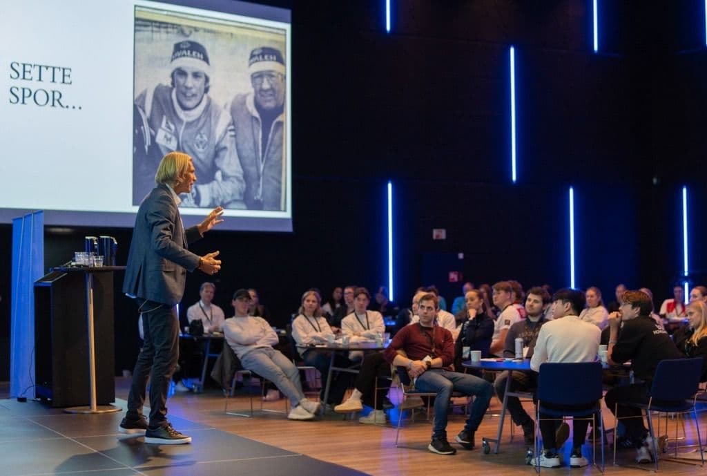 En foredragsholder står på en scene foran et publikum av studenter, med teksten Sette spor og et bilde av to skiløpere på lerretet.