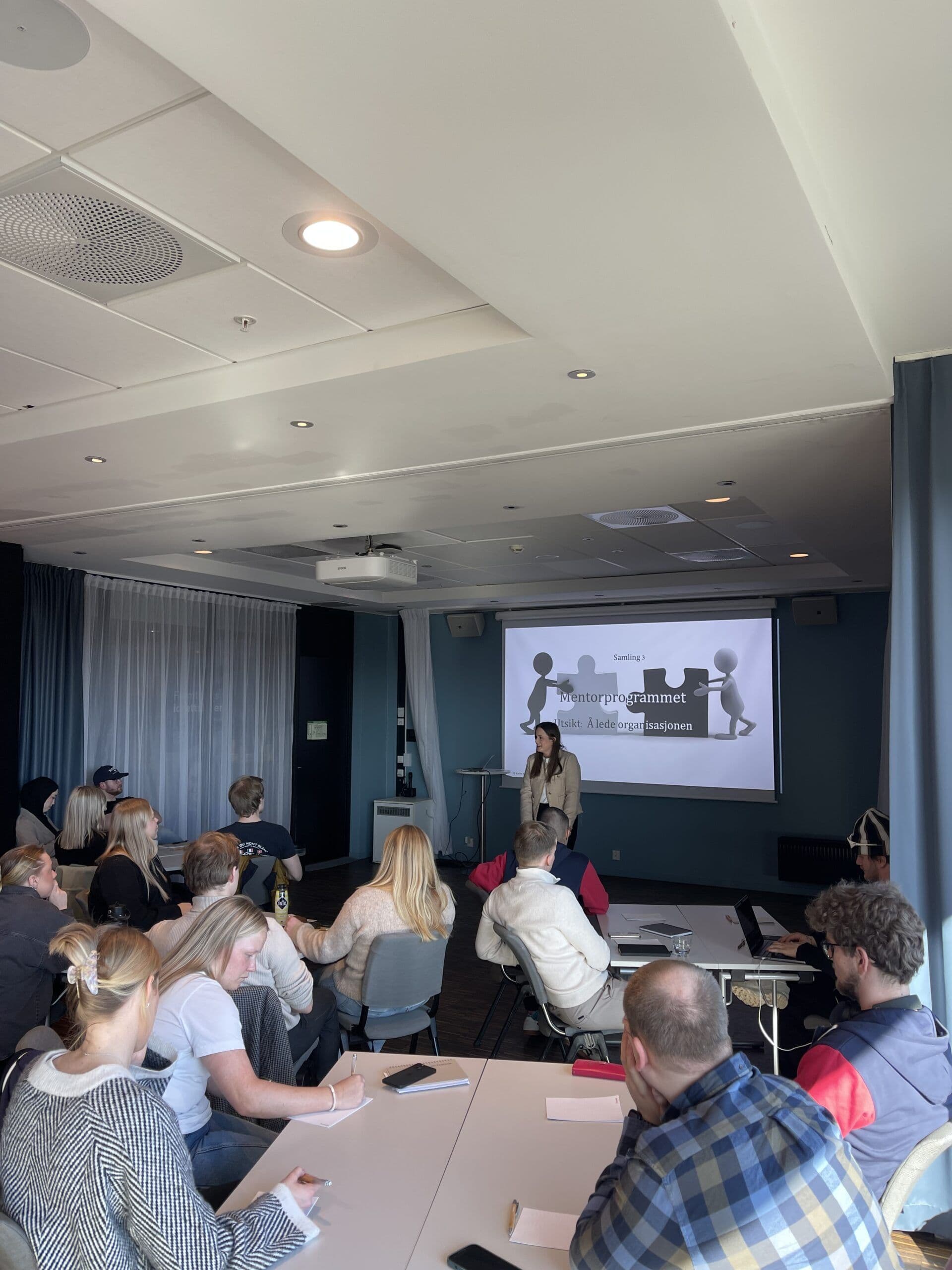 En gruppe deltakere sitter og følger med på et foredrag om mentorprogrammet i et møterom med presentasjon på lerretet.